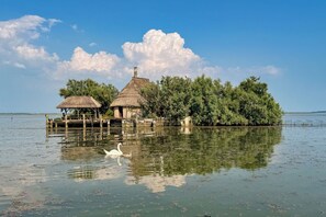 Ein Gebäude mit strohgedecktem Dach, im Vordergrund ein Gewässer und im Hintergrund ein bewaldetes Gebiet.