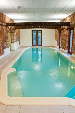 Una piscina interior con fondo de pared de ladrillo, una pérgola de madera y suelo de baldosas.