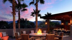 A round outdoor seating area with a fire pit, surrounded by wooden chairs and tables, set against a backdrop of palm trees and mountains under a twilight sky. - Luxurious Resort Stay, 2br lock off at Westin Palm Desert (Palm Desert)