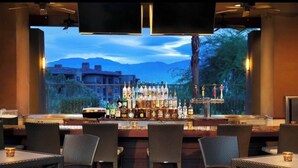 A bar area with wicker chairs and tables, a variety of bottles, and a view of mountains through large windows. - Luxurious Resort Stay, 2br lock off at Westin Palm Desert (Palm Desert)
