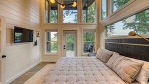 A bedroom with a large bed, a TV, and a view of the outdoors through multiple windows. - Mountain Abode - Hot tub & mountain views (Blue Ridge Mountains)