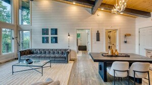 A spacious living area with a large sofa, a coffee table, and a dining area with chairs. There are large windows providing natural light and a view of the outdoors. - Mountain Abode - Hot tub & mountain views (Blue Ridge Mountains)