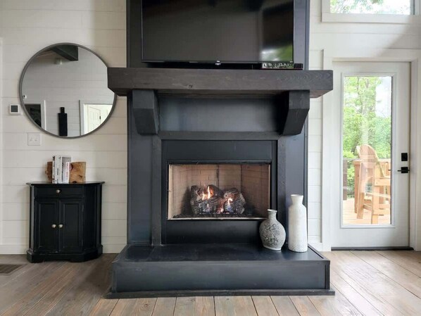 A modern fireplace with a TV above it, a round mirror on the wall, and a small cabinet with books. - Mountain Abode - Hot tub & mountain views (Blue Ridge Mountains)