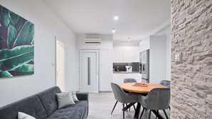 Een moderne woonkamer met een grijze bank, een houten eettafel met stoelen, en een keuken met een koelkast en magnetron.