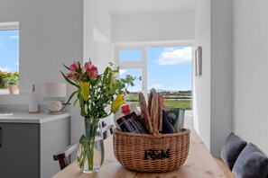 Un coin repas offrant une vue dégagée sur l’extérieur, un panier en osier rempli de magazines et un vase avec des fleurs posé sur une table.