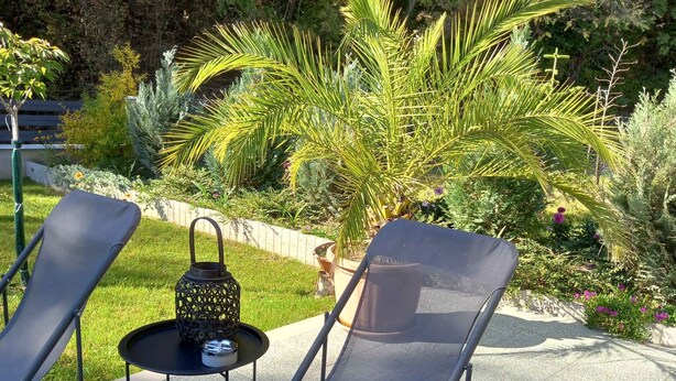 Dos sillones de exterior con una mesita y un farol decorativo, ubicados en un patio empedrado con un jardín al fondo.