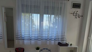 Una habitación con un ventanal grande cubierto por cortinas translúcidas, una mesita consola blanca con una planta, un estante pequeño y un espejo en la pared.