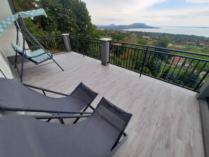 A balcony with two lounge chairs and a table, overlooking a scenic landscape with trees and a distant view of a body of water and mountains. - Holiday home with view and pool (Balatongyörök)
