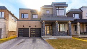 A modern two-story residential building with a garage, a covered front porch, and a well-maintained lawn. - Lakeside Modern House 4BR - Steps to the lake (Bowmanville)
