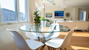 A modern dining area with a glass table and white chairs, a potted plant, and a view of the city through large windows. - Lakeside Modern House 4BR - Steps to the lake (Bowmanville)