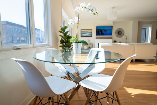 A modern dining area with a glass table and white chairs, a potted plant, and a view of the city through large windows.