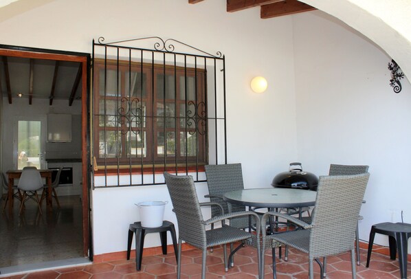 Un patio al aire libre con sillas de mimbre, una mesa redonda, una parrilla y vistas del interior a través de una ventana.