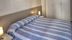 A bedroom with a bed, a nightstand, a lamp, a wardrobe, and a wall decoration. - Holiday apartment with balcony and air conditioning (Lignano Sabbiadoro)