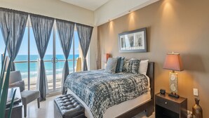 A bedroom with a large bed, a ceiling fan, a nightstand, a lamp, and a view of the ocean.