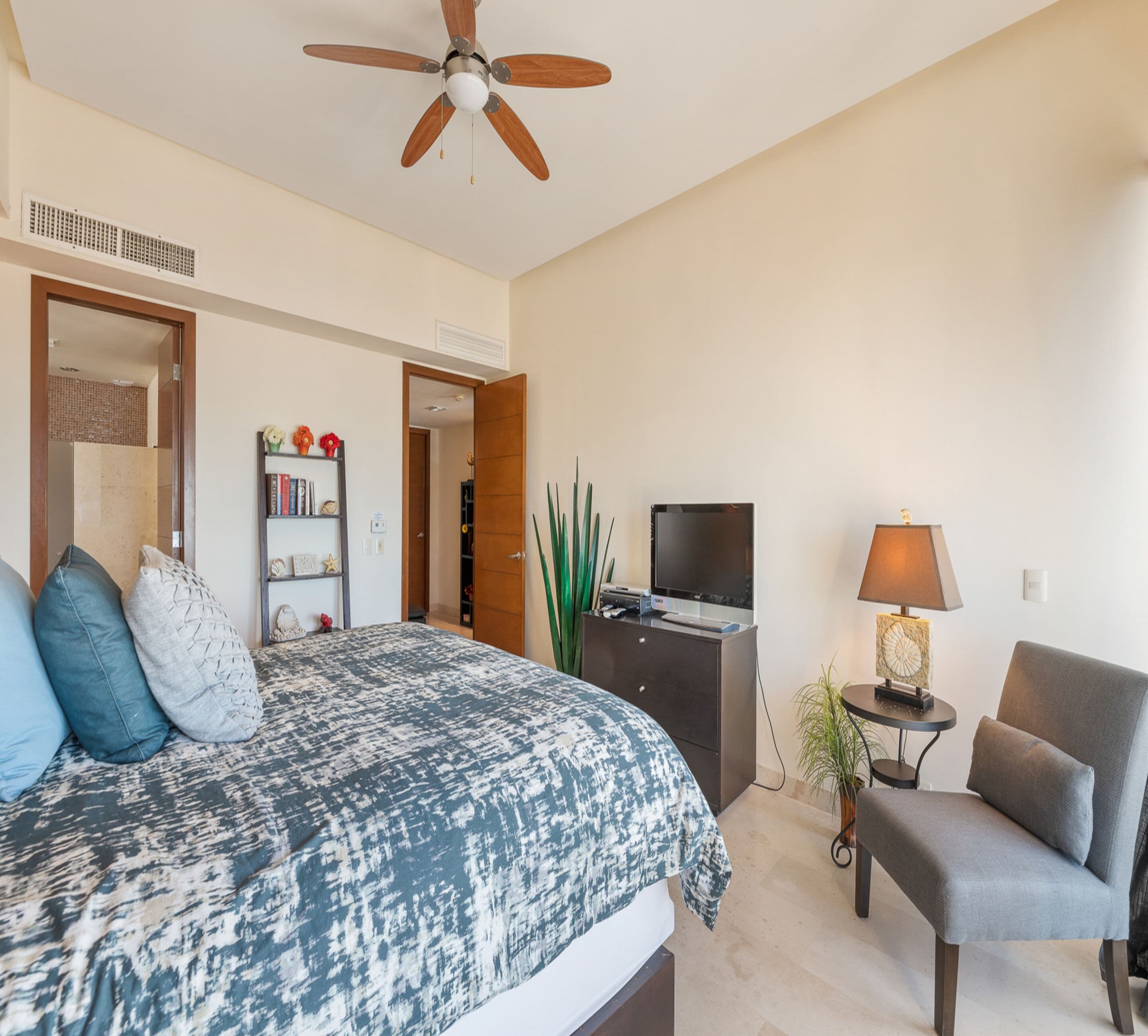 A bedroom with a bed, a TV, a chair, a lamp, and a ceiling fan.