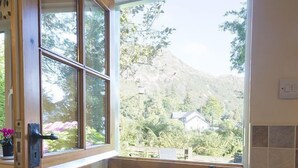 A kitchen with a window overlooking a mountainous area, wooden cabinets, and a tiled backsplash. - Yewdale Cottage, village home with mountain views (Coniston)