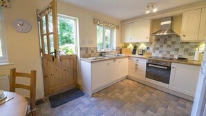 A compact kitchen with white cabinets, a stove, and a window overlooking greenery. - Yewdale Cottage, village home with mountain views (Coniston)