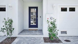 Une maison moderne avec une porte d’entrée bleu foncé, des portes de garage blanches et un petit porche à l’avant, agrémenté de deux plantes en pot.