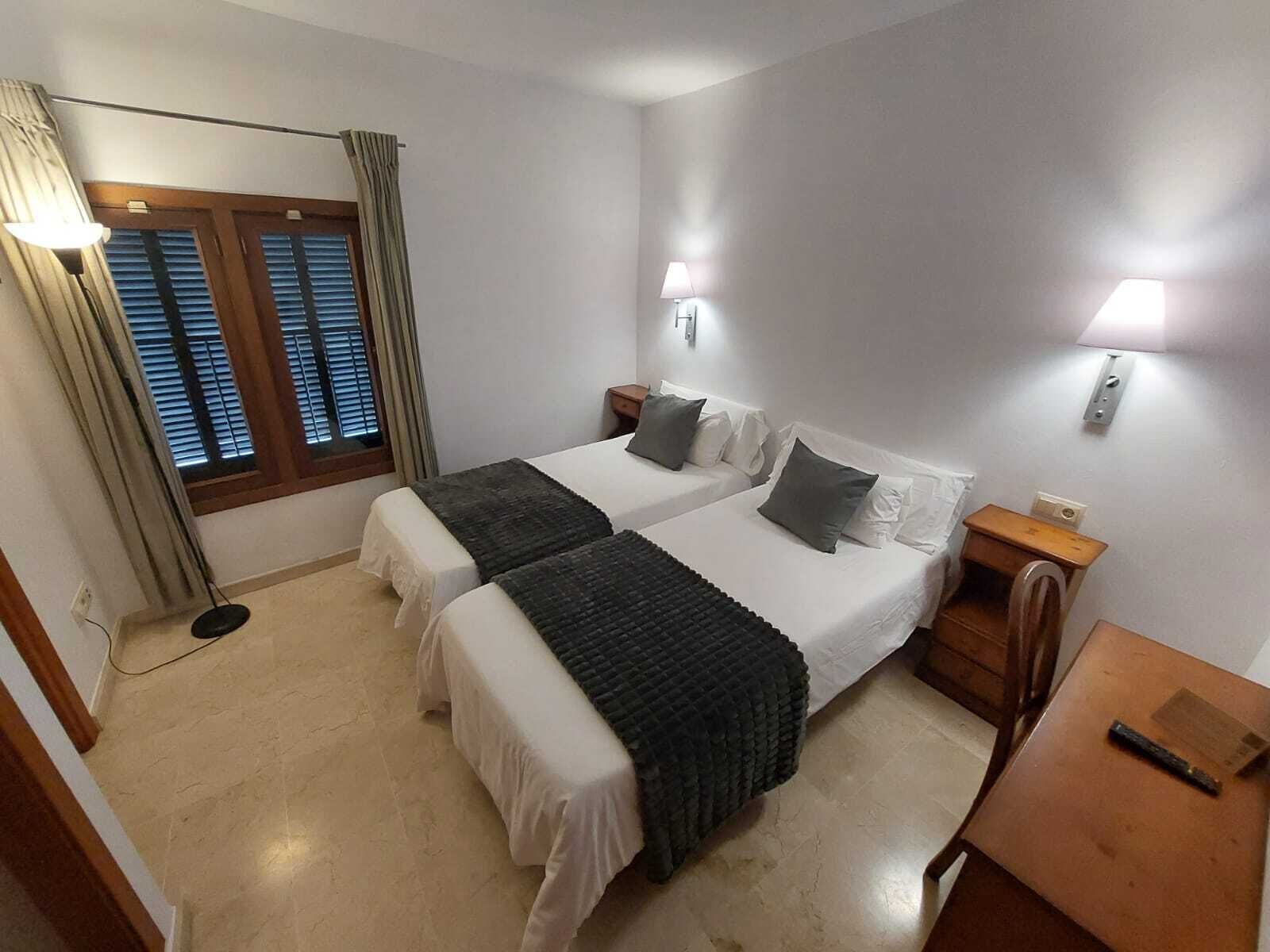 Habitación de hotel con dos camas, un escritorio, una silla, una lámpara y una ventana con persianas.