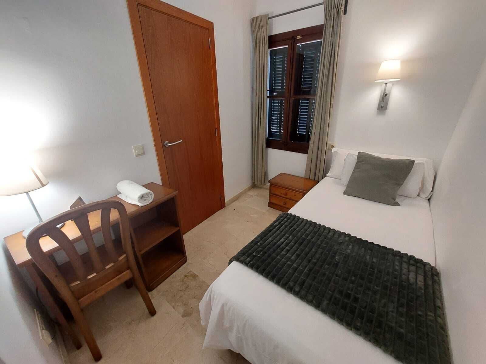 Un dormitorio con cama, escritorio, silla, mesita de noche y ventana con cortinas.
