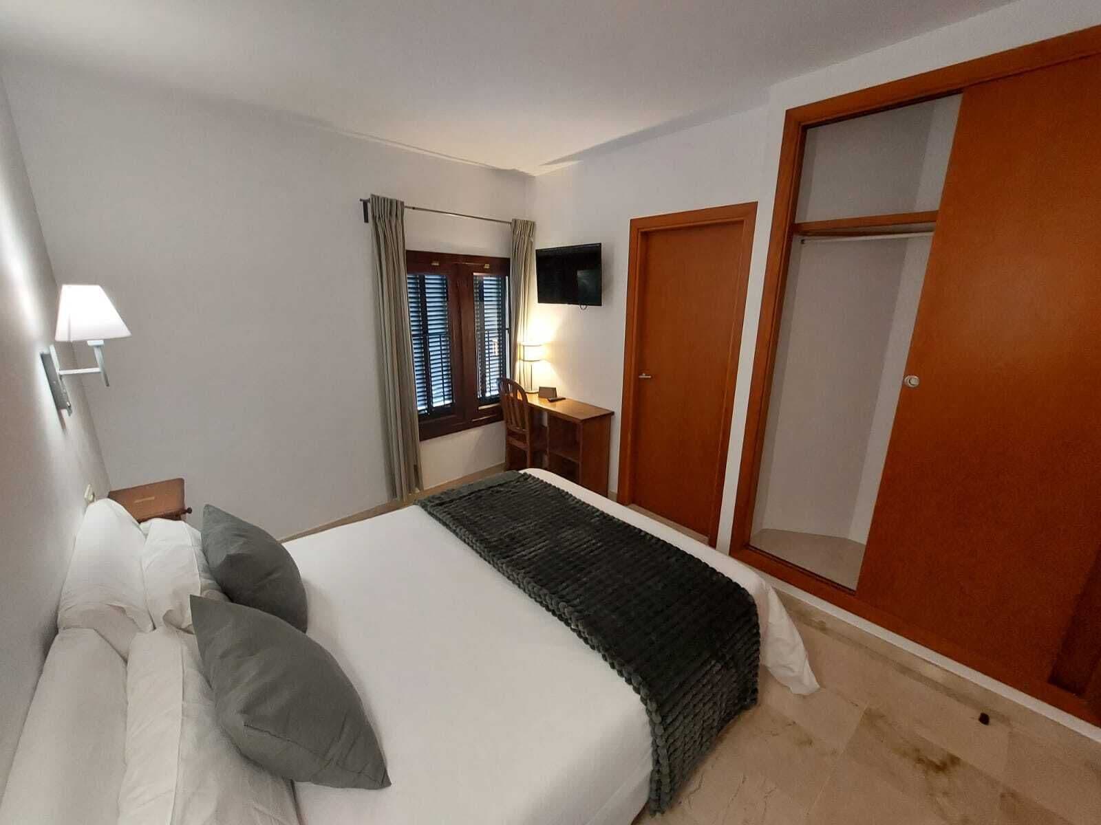 A hotel room with a bed, bedside table, lamp, window with curtains, and a wardrobe.