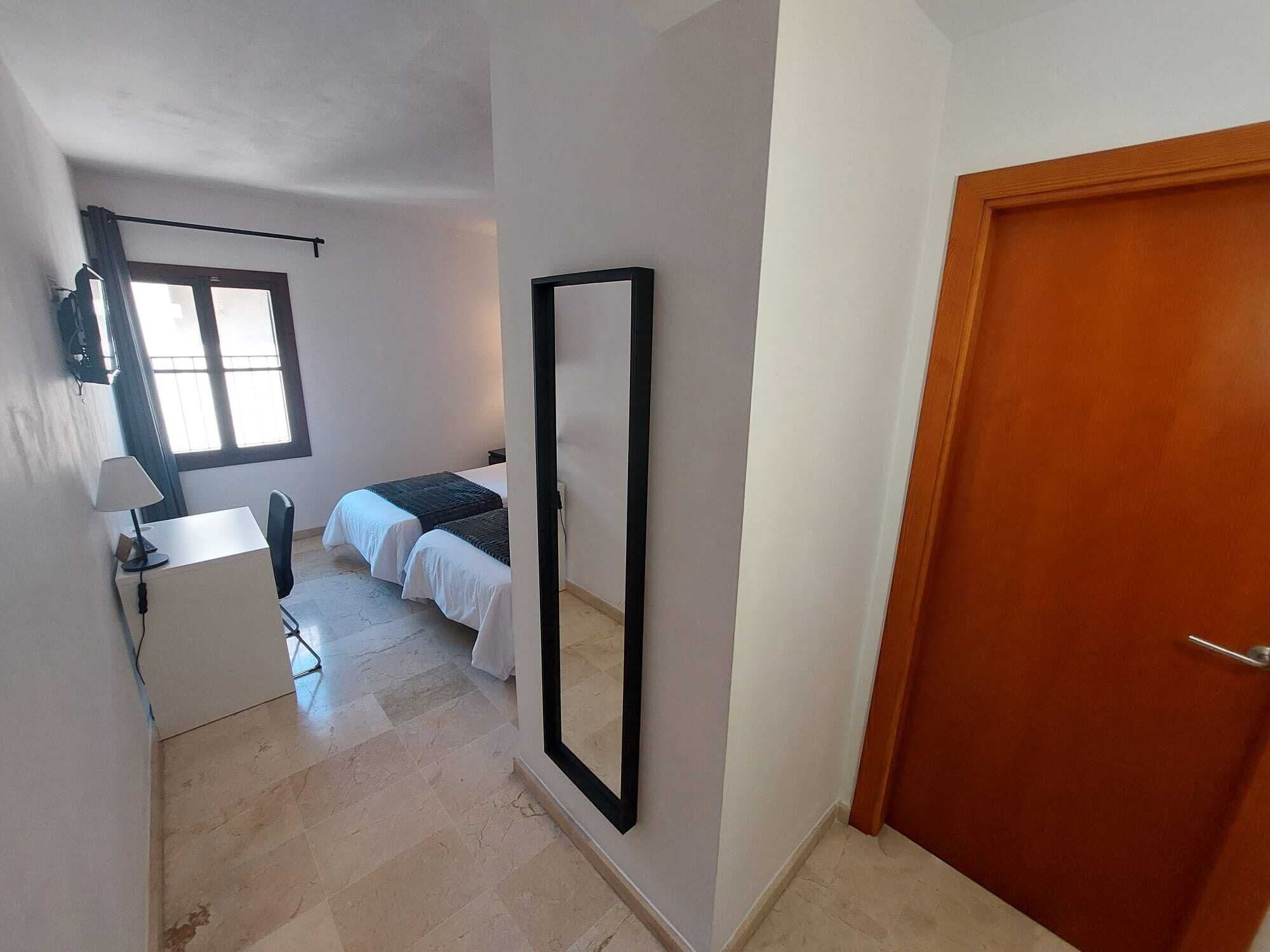 Een slaapkamer met een bed, een bureau met een lamp, een grote spiegel en een deur.