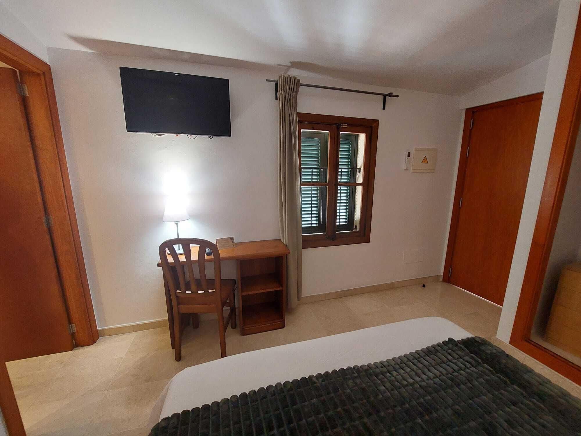 A room with a bed, a desk with a chair, a television mounted on the wall, a window with curtains, and a door.