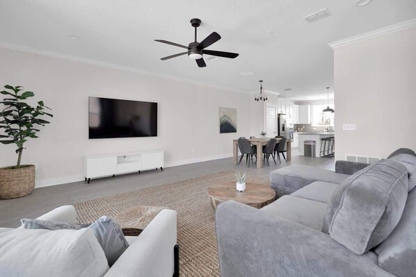 A modern living room with a flat-screen TV, grey sofas, a ceiling fan, and a dining area in the background.