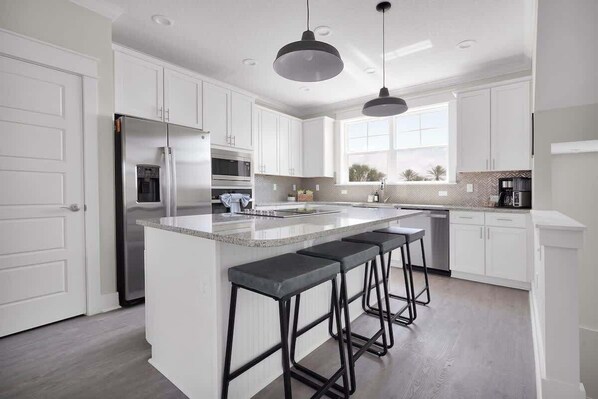 A modern kitchen with white cabinetry, a stainless steel refrigerator, a built-in oven, a large island with bar stools, pendant lighting, and a window providing natural light.