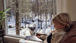 Une personne est assise près d’une fenêtre, en train de lire un magazine, et la vue donne sur une forêt enneigée.