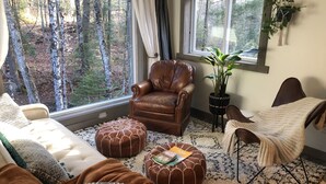Un salon chaleureux avec un fauteuil en cuir, un pouf et une vue sur une forêt, visible à travers de grandes fenêtres.