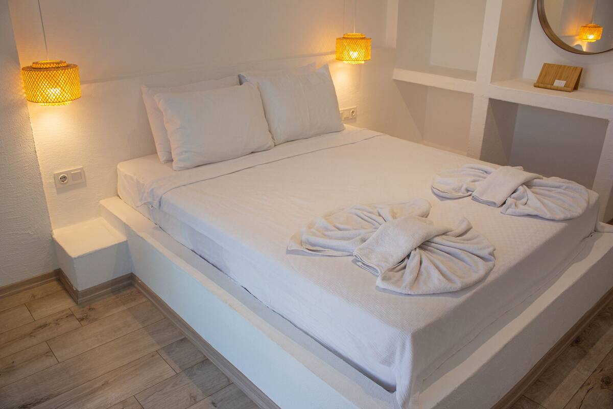Uma cama bem arrumada com lençóis e toalhas brancas, duas luminárias na parede e uma pequena mesa de cabeceira.
