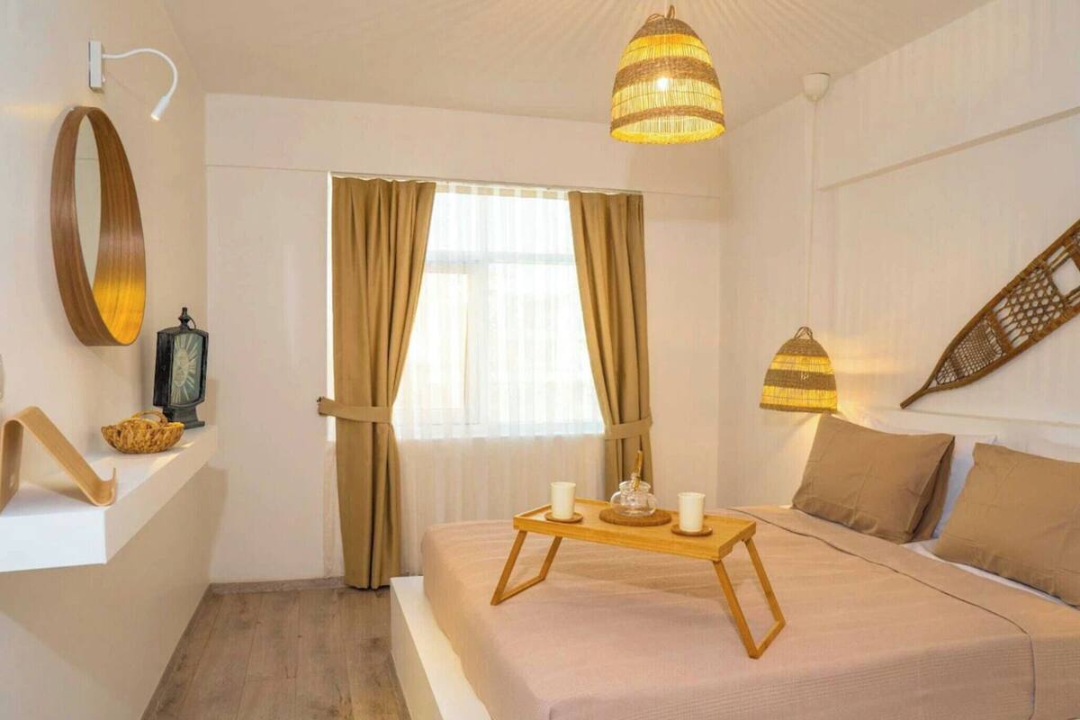 Quarto de hotel com cama, mesa de cabeceira com bandeja, luminária pendente, espelho fixado na parede e janela com cortinas.