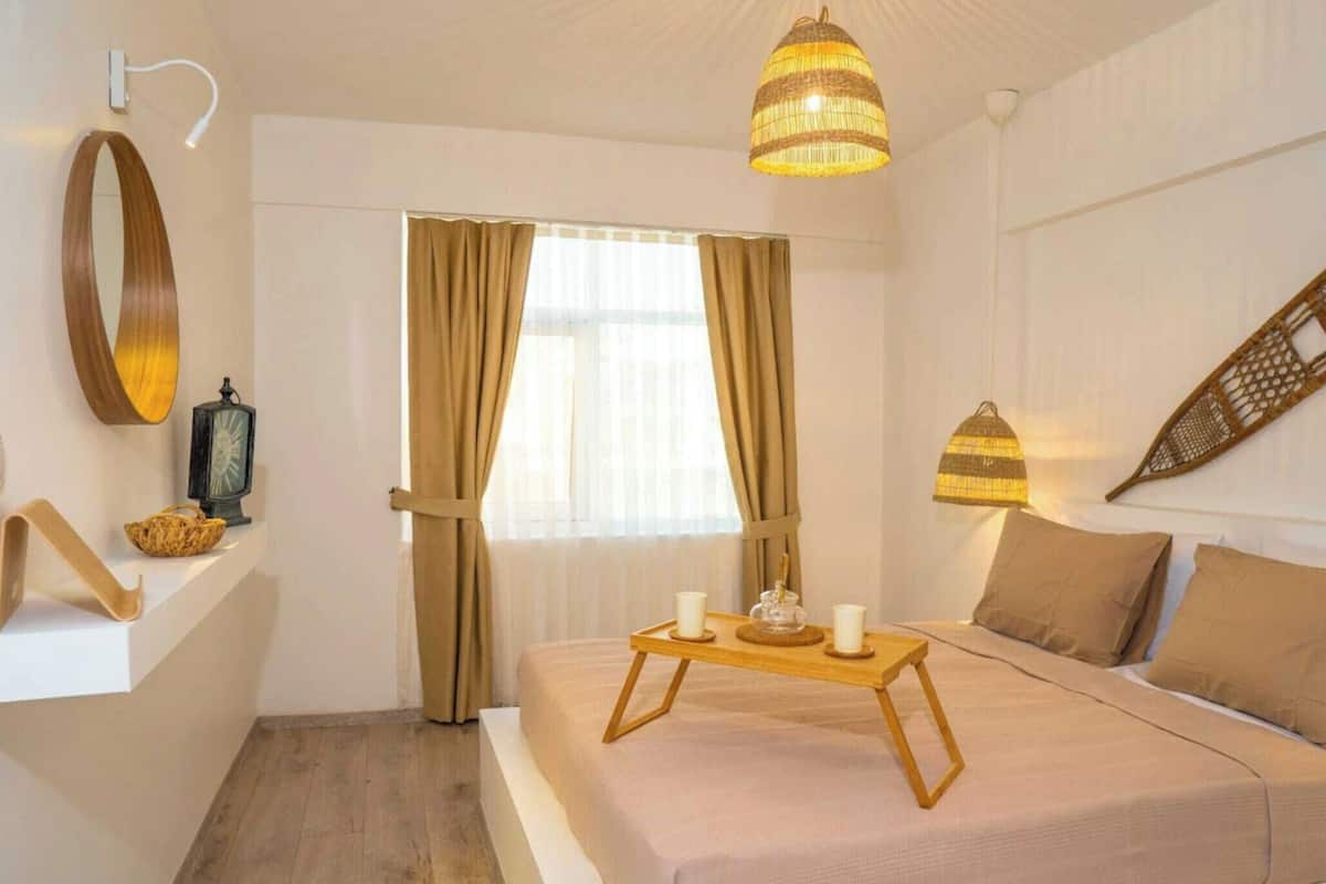 A hotel room with a bed, a bedside table with a tray, a hanging lamp, a wall-mounted mirror, and a window with curtains.