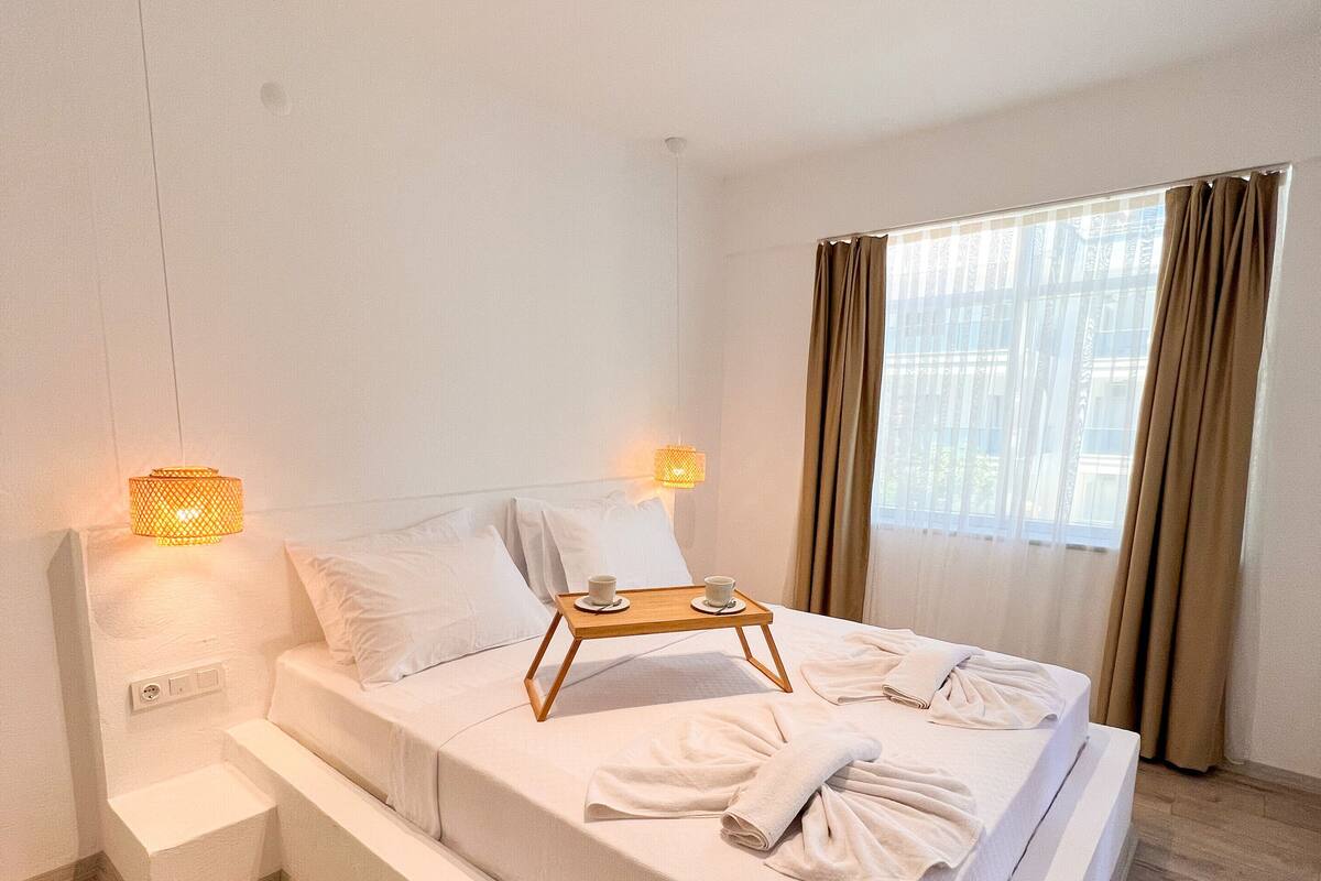 Quarto de hotel moderno com uma cama grande, luminárias de cabeceira, mesa com xícaras e vista para a cidade pela janela.