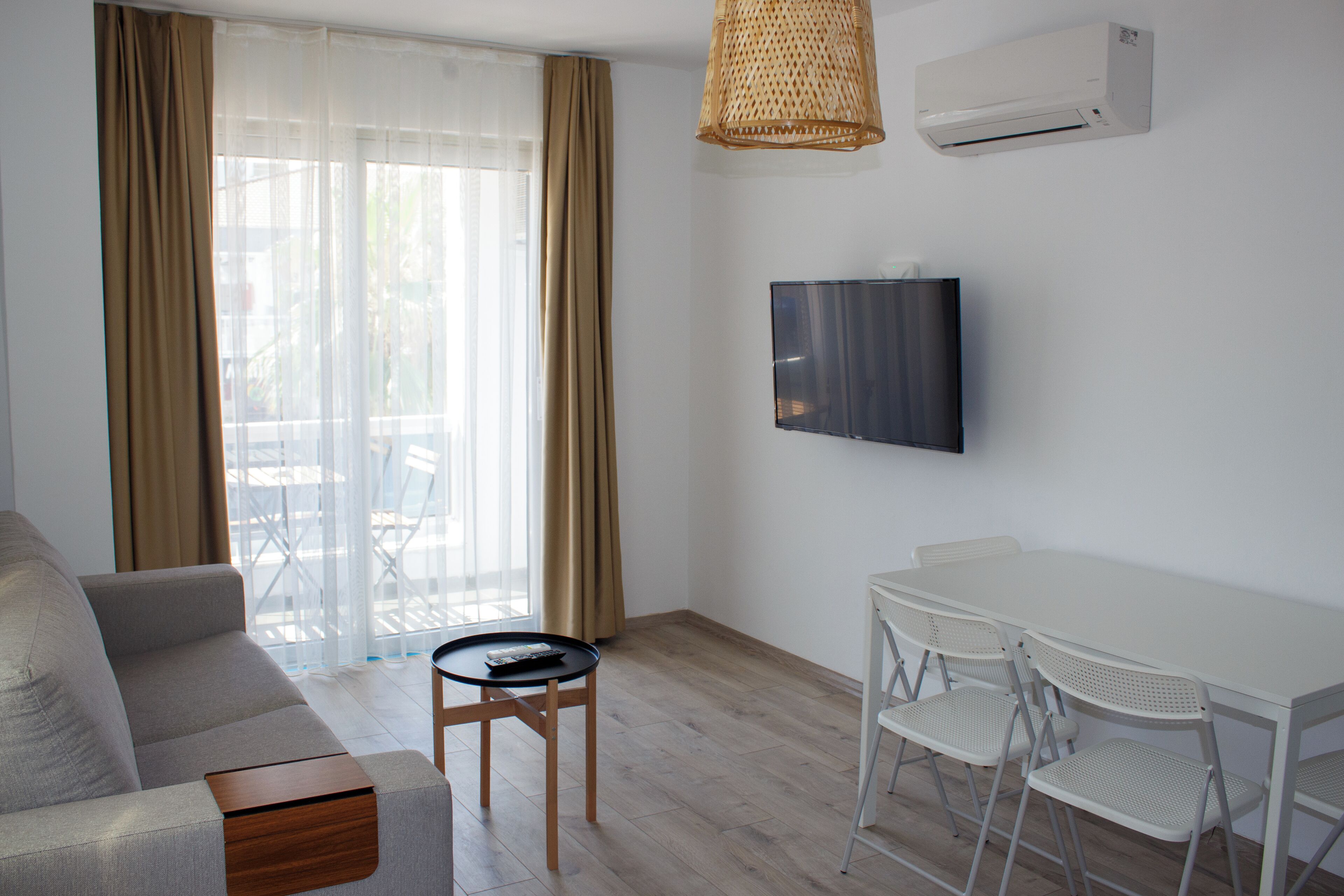 A modern living room with a sofa, a small table, a TV mounted on the wall, and a dining area with chairs.