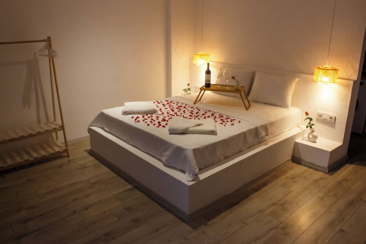 Cama bem arrumada com colcha branca, uma pequena mesa de cabeceira com uma garrafa e copos, e uma luminária fixada na parede.
