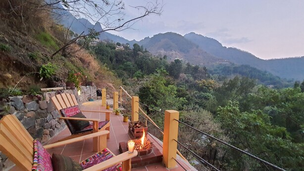 A terrace with wooden chairs and a fire pit, overlooking a mountainous landscape.