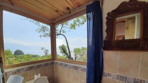 A bathroom with a toilet, sink, and a window offering a view of greenery and a distant tree.