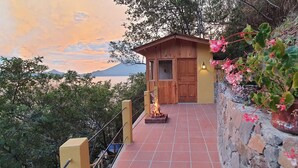 A wooden cabin with a stone wall and a fire pit, overlooking a body of water and mountains.