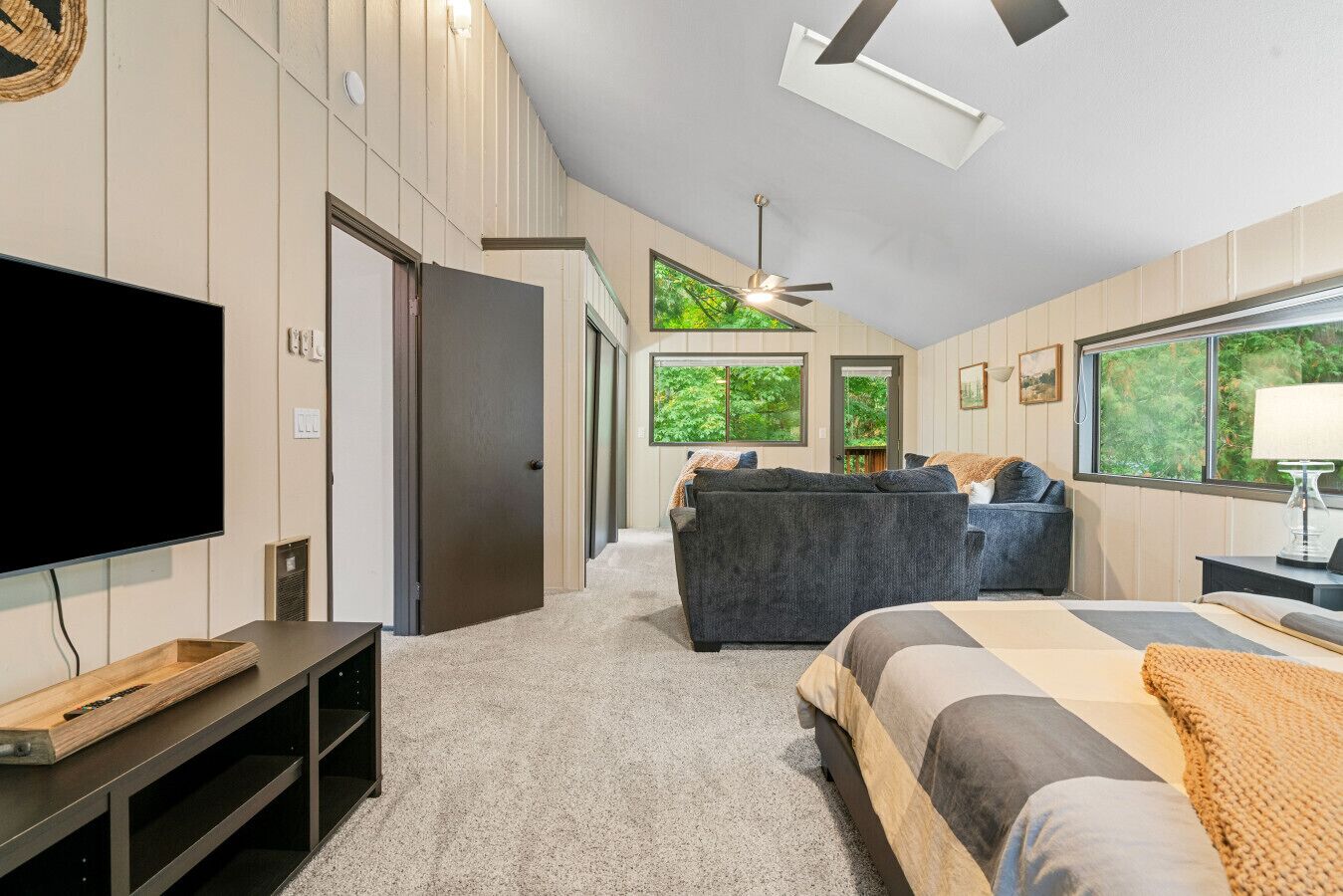 Une chambre à coucher moderne, équipée d’un lit, d’un téléviseur, d’un ventilateur de plafond et offrant une vue sur la verdure par les fenêtres.