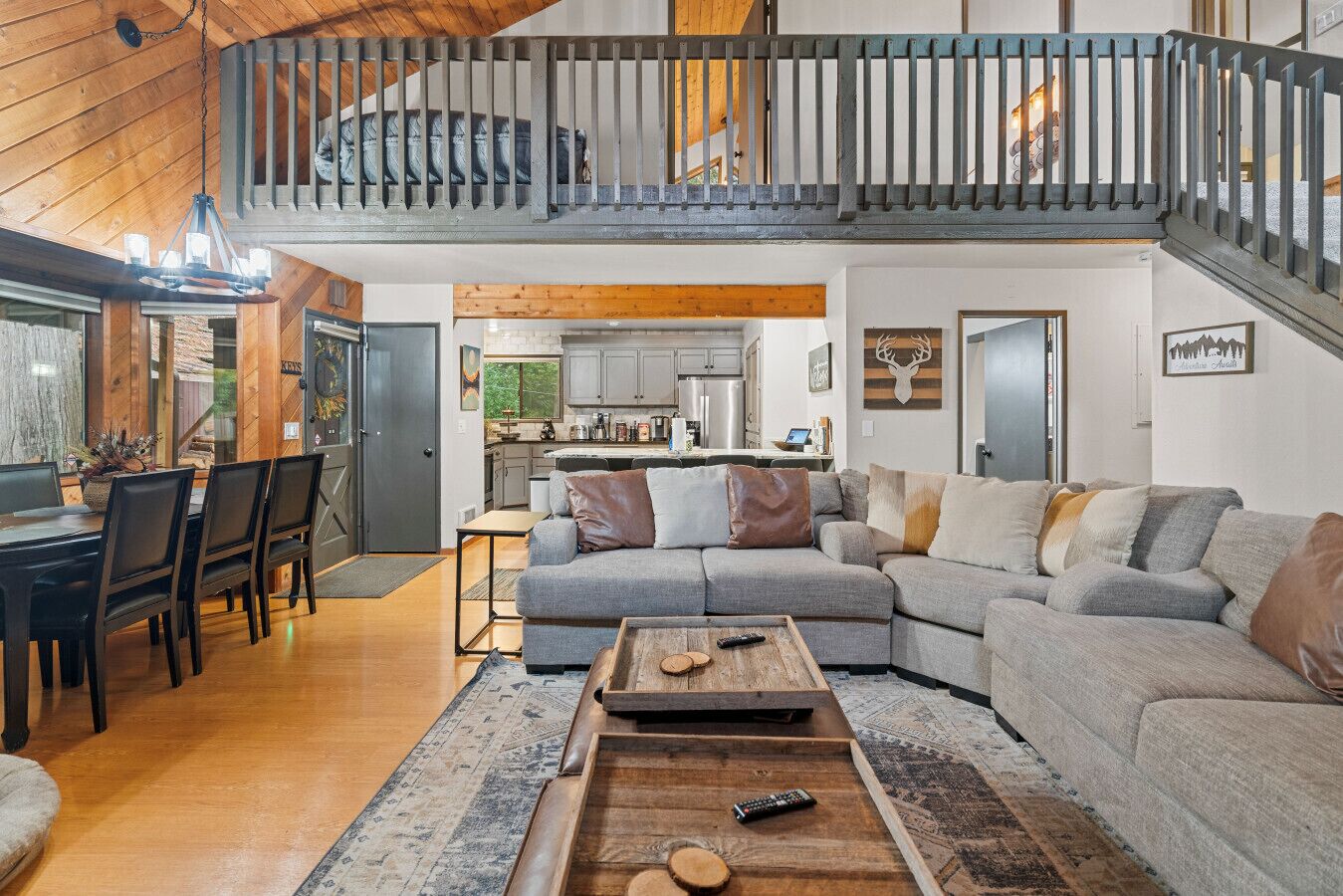 A spacious living room with a grey sectional sofa, wooden coffee table, and a dining area with a dark wooden table and chairs. A kitchen is visible in the background with modern appliances.