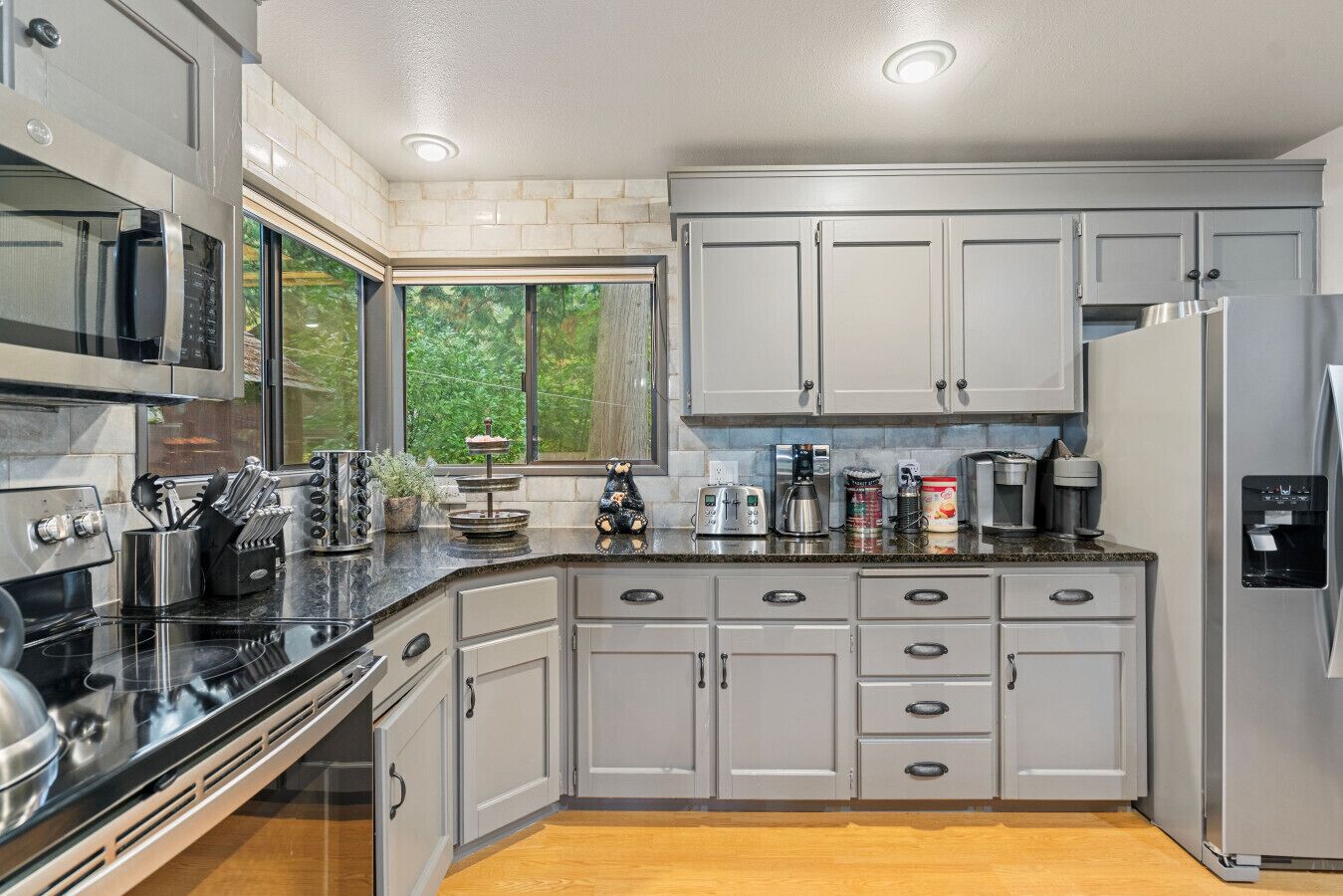 A modern kitchen with grey cabinets, stainless steel appliances, and a window providing a view of greenery outside.