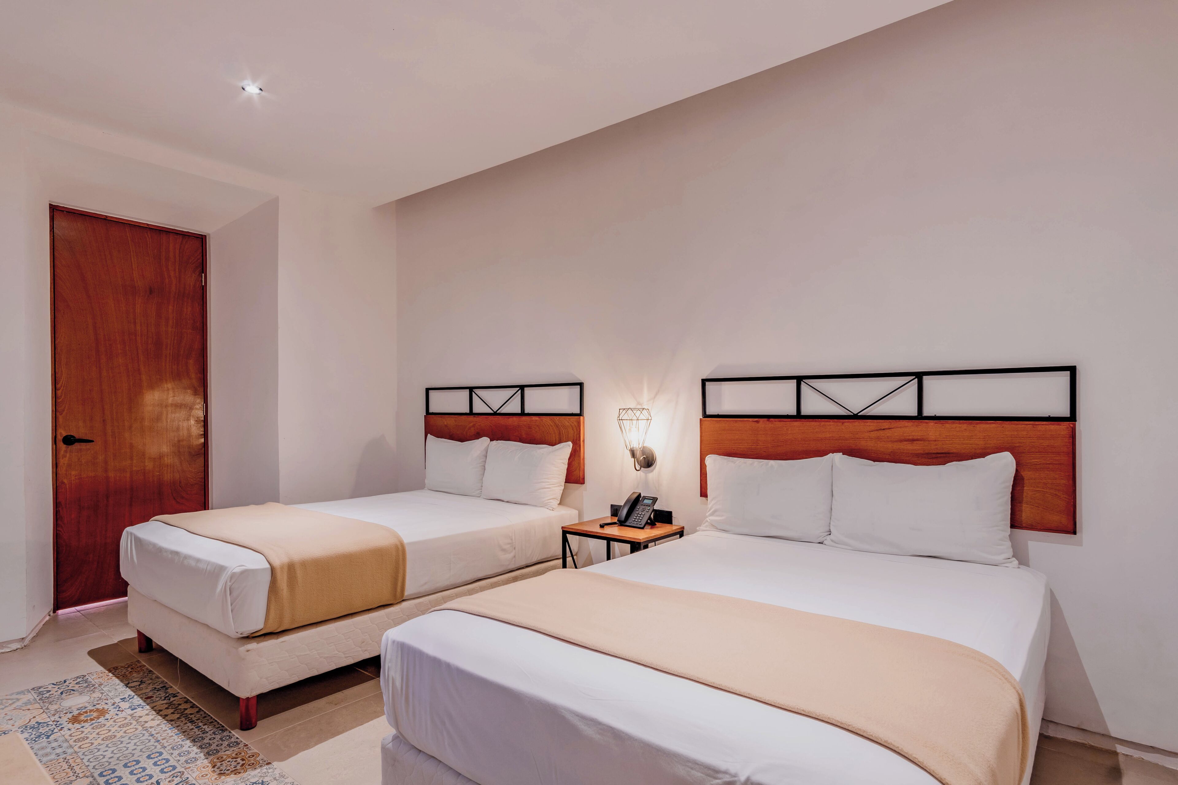 Habitación de hotel con dos camas, una puerta de madera, una mesita de noche con lámpara y un tapete estampado.