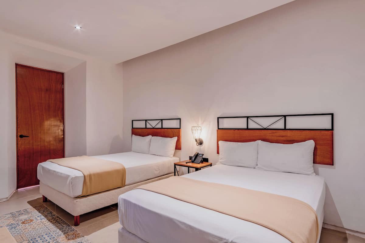 Habitación de hotel con dos camas, una puerta de madera, una mesita de noche con lámpara y un tapete estampado.