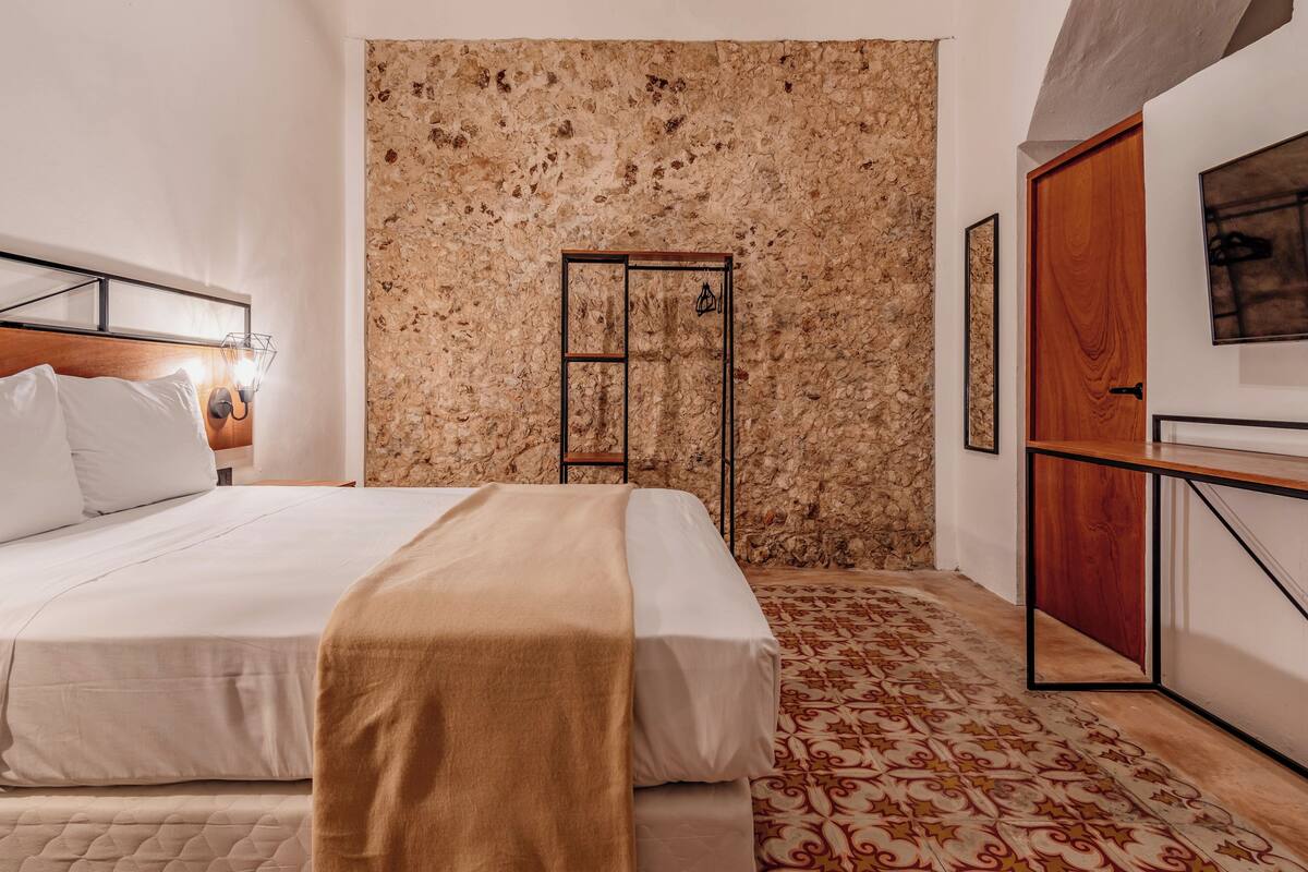 Ett sovrum med en säng, en trädörr, en stenmur och en mönstrad matta.