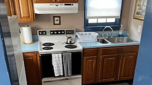 A compact kitchen with wooden cabinets, a white stove, a sink, and a window with blinds. - Waterfront Cottage on the C&D Canal! (Chesapeake City)