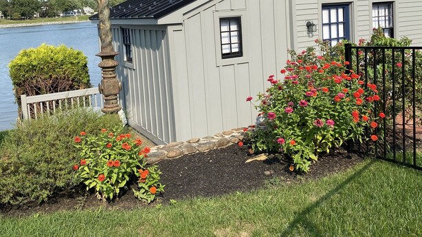 A two-story house with a light blue exterior, a black roof, and a white fence. There is a lamp post and flowering plants in the front yard. - Waterfront Cottage on the C&D Canal! (Chesapeake City)