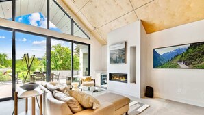 A modern living room with a sofa, coffee table, and fireplace, large windows, and a mountain view.