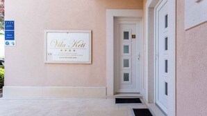 L’entrée d’un hôtel, avec une enseigne encadrée portant l’inscription « Vila Keti », une porte avec une plaque numérotée et un panneau bleu indiquant les services proposés.